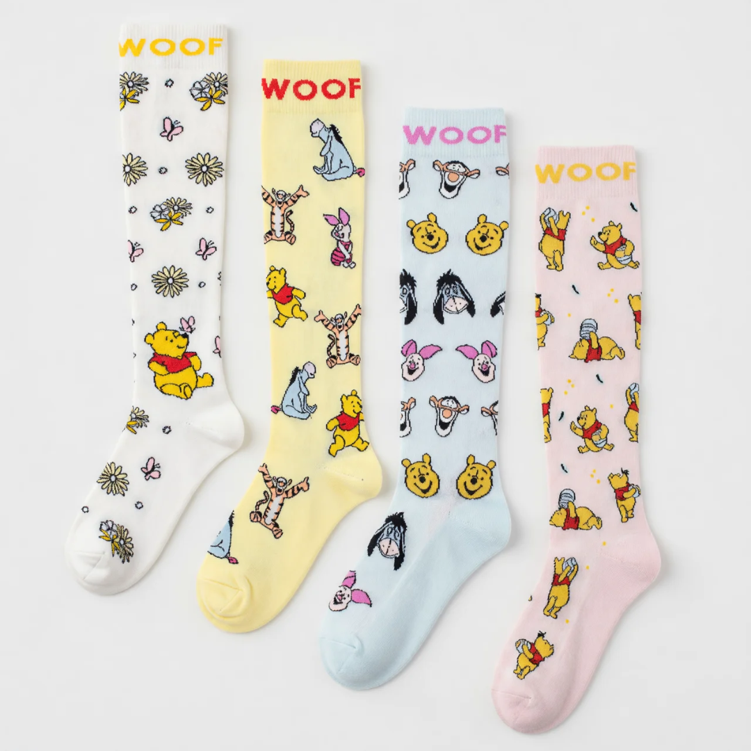 Studio layout displaying the Disney’s Winnie the Pooh Bamboo Compression Socks 4 Pack laid out side by side, featuring four distinct pastels: white floral Pooh print, pale yellow Hundred Acre Wood character print, light blue character face print, and soft pink hunny pot Pooh print—each styled with contrast "WOOF" top cuff branding.   