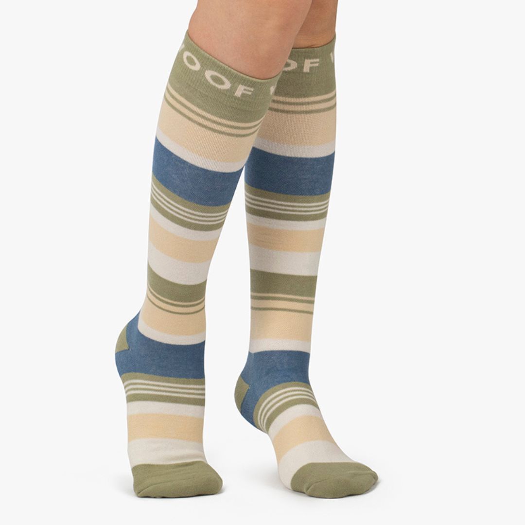 Nurse wearing knee-high compression socks featuring horizontal stripes in olive green, cream, and slate blue, finished with top cuff branding.